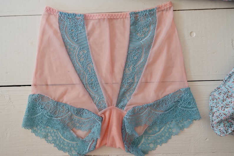 May include: A pair of pink underwear with teal lace trim. The underwear features a sheer pink fabric with teal lace accents along the edges and down the front. The lace has a floral design.