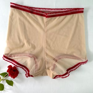 Puede incluir: Ropa interior beige transparente con ribete rojo en la cintura y las aberturas de las piernas. La ropa interior de cintura alta est&aacute; hecha de una tela delicada y transparente. Una rosa roja es visible en la esquina inferior izquierda.