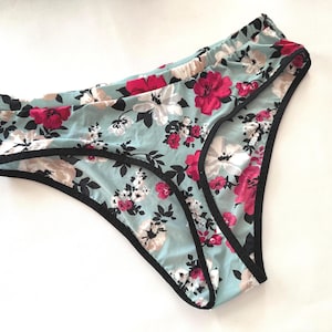 May include: Pale blue underwear with a floral print of pink and white flowers, edged with black trim. The low-rise briefs are made from a soft, lightweight material. The floral pattern is consistent across the garment.
