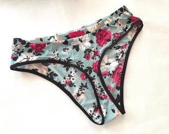 Op de afbeelding: Lichtblauwe onderbroek met een bloemenpatroon met roze en witte bloemen en zwarte rand. De onderbroek heeft een lage taille en is gemaakt van een zachte, lichte stof. Het bloemmotief herhaalt zich over het hele kledingstuk.