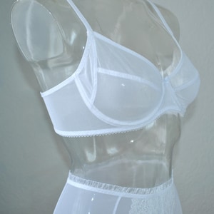 Op de afbeelding: Witte, doorschijnende bh en slipset met delicate kantversiering. De bh heeft een klassieke beugel en verstelbare bandjes. De slip heeft een hoge taille en een kantversiering rond de beenopeningen.