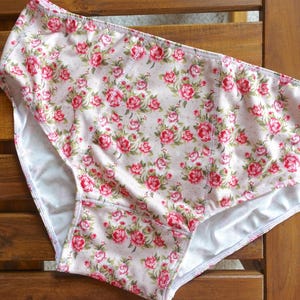 May include: High-waisted knickers featuring a light pink background with a repeating pattern of small red roses and green leaves. The garment has a white interior lining and visible stitching along the seams, providing a full-coverage design.