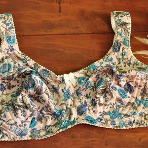 Pode incluir: Um sutiã branco com um padrão floral em tons de azul e rosa. O sutiã tem uma borda de renda branca e um fecho de gancho e olho nas costas.