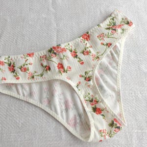 Könnte beinhalten: Eine gerippte Unterhose in Off-White mit einem wiederkehrenden Blumenmuster aus kleinen roten Rosen und grünen Blättern. Das Kleidungsstück hat einen hoch geschnittenen Beinausschnitt und einen breiten Bund aus weichem, strukturiertem Stoff.