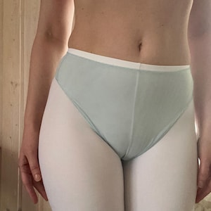 Op de afbeelding: Lichtblauwe high-waisted onderbroek met een witte tailleband. Het ondergoed wordt gecombineerd met witte leggings. Een roze bh is zichtbaar op de achtergrond. De foto is binnenshuis gemaakt.