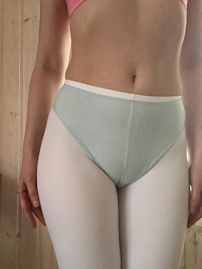 Op de afbeelding: Lichtblauwe high-waisted onderbroek met een witte tailleband. Het ondergoed wordt gecombineerd met witte leggings. Een roze bh is zichtbaar op de achtergrond. De foto is binnenshuis gemaakt.