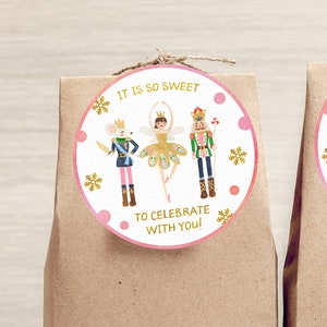 Puede incluir: Dos bolsas de regalo de papel marrón con pegatinas redondas. Las pegatinas tienen un borde rosa y presentan ilustraciones de un ratón, una bailarina y un cascanueces. El texto de la pegatina dice "IT IS SO SWEET TO CELEBRATE WITH YOU!"