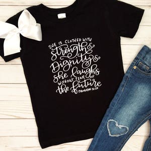 May include: Black t-shirt with white script reading "She is clothed with Strength & Dignity & she laughs without fear of the future Proverbs 31:25." A white bow and blue jeans with a heart-shaped patch are also visible.