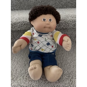 May include: A vintage doll with a soft body and painted face. The doll has brown hair, a white sweater with a geometric pattern, blue corduroy trousers, and large, beige hands and feet. The doll is sitting on a grey carpet.