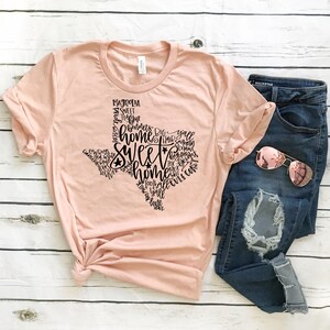 May include: A light pink t-shirt with a black Texas-shaped design filled with words like "home" and "sweet home." The shirt is paired with blue jeans and rose-tinted sunglasses, all arranged on a white wooden surface.