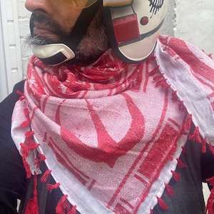 Puede incluir: Un pañuelo con estampado rojo y blanco con borlas rojas. El pañuelo está drapeado sobre una chaqueta oscura. Un casco blanco con detalles en rojo y negro es visible en el fondo.