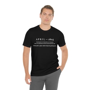 May include: Black t-shirt with white text. The text reads "APRIL - 1805, NAPOLEON IS MASTER OF EUROPE, ONLY THE BRITISH FLEET STANDS BEFORE HIM, OCEANS ARE NOW BATTLEFIELDS." The shirt is a crew neck style.