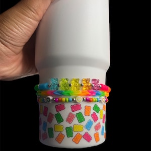 May include: White tumbler with a colorful gummy bear design. The tumbler has three bands of colorful beads and gummy bears around the top.