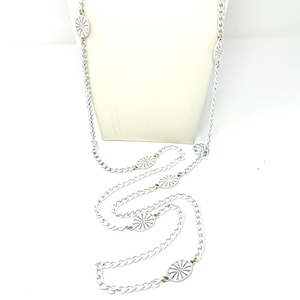 Op de afbeelding: Een lange, zilverkleurige ketting met een delicaat kettingontwerp. De ketting heeft verschillende cirkelvormige, wit en zilverkleurige decoratieve elementen. De ketting wordt getoond op een witte mannequin, die de lengte en het ontwerp benadrukt.