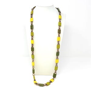 May include: A long necklace with rectangular links in shades of yellow and green, connected by bronze-colored metal. The necklace is displayed on a white mannequin, showcasing its length and design. The color palette is earthy and vibrant.