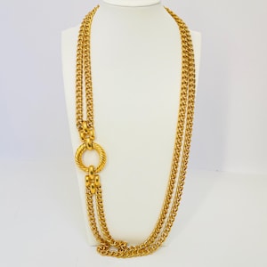 Puede incluir: Un collar de cadena de oro con un gran colgante redondo de oro. El collar está en capas y tiene una cadena gruesa y texturizada.