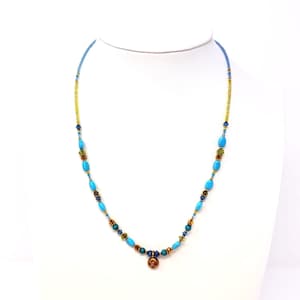 May include: A beaded necklace with turquoise, gold, and dark blue beads. The necklace has a small, round, copper-colored pendant. The necklace is displayed on a white mannequin bust.