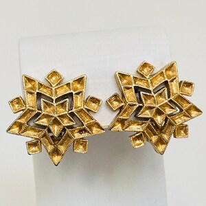 May include: A pair of gold-toned, geometric snowflake earrings. Each earring features a multi-faceted design with a central starburst pattern and pointed edges. The earrings are displayed on a white stand against a white background.