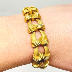 May include: A gold-toned bracelet with a woven design. The bracelet features a series of interconnected, textured links, creating a bold and intricate pattern. The warm gold color gives the bracelet a classic and elegant look. The bracelet is approximately 7 inches in length.