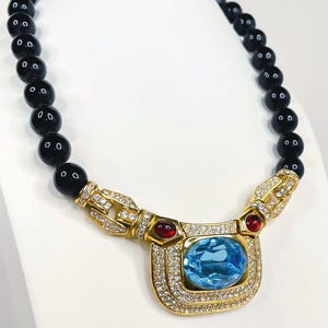 Può includere: Collana dorata con una grande gemma rettangolare azzurra al centro, circondata da pietre chiare più piccole. La collana ha una catena di perline nere e dettagli rossi su entrambi i lati della pietra centrale. Esposta su un busto bianco.