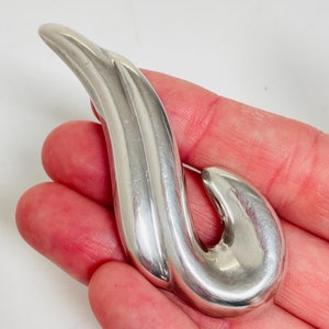 May include: A silver abstract brooch with a curved, flowing design. The brooch has a smooth, polished finish.