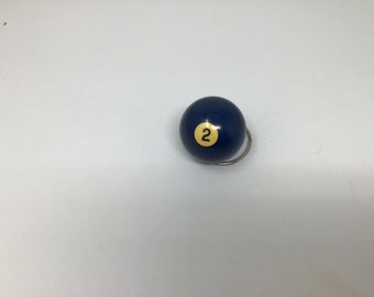May include: A blue billiard ball with a yellow number 2 on it. The ball is attached to a silver key ring.