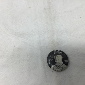 May include: Black and white button with a portrait of a man in a hat. The text "B.P. 1857 1907 1957" is printed around the portrait.