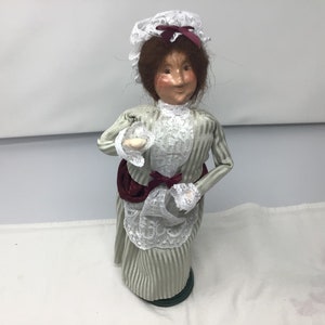 May include: A vintage-style doll wearing a white lace dress with green and white stripes, a white lace bonnet, and a burgundy sash. The doll is holding a small white object in her right hand.