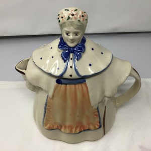 May include: A ceramic teapot shaped like a woman wearing a white dress, blue shawl, and a floral crown. The teapot has a brown handle and a spout.
