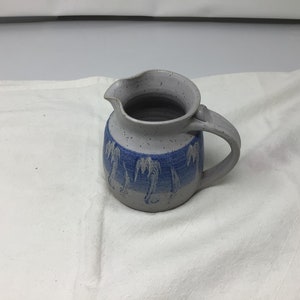 May include: A gray ceramic pitcher with a blue and white design of palm trees. The pitcher has a speckled glaze and a rounded body.