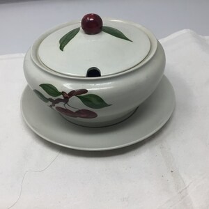 May include: White ceramic serving dish with a lid, featuring a cherry motif with green leaves and red cherries. The dish has a small spout on the lid.