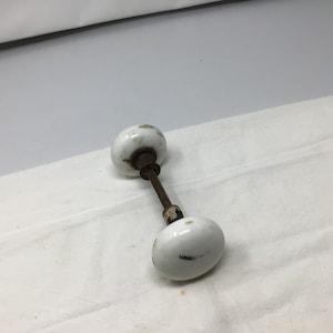 May include: A pair of white porcelain door knobs with a metal spindle connecting them. The knobs have a slightly aged look with some discoloration.