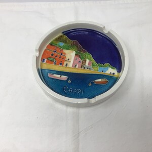 May include: White ceramic ashtray with a colorful painted scene of a town on a hillside with a blue sea and two boats. The text "CAPRI" is written on the bottom of the ashtray.
