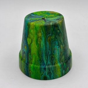 May include: A small, painted terracotta flower pot with a vibrant, swirled design. The color palette includes shades of green, blue, and gold, creating a marbled effect. The pot has a small drainage hole at the bottom and is suitable for small plants.