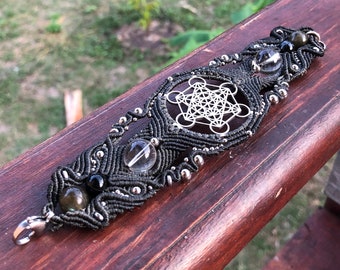 May include: A black macrame bracelet with silver beads and a silver metal geometric design. The bracelet has clear and green stones.