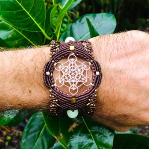 Puede incluir: Un brazalete de macramé marrón con un diseño geométrico de metal blanco y cuentas doradas. El brazalete se lleva en la muñeca.