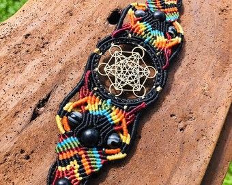 Puede incluir: Pulsera de macramé negra con un dije geométrico dorado y cuentas negras. La pulsera está tejida con hilos de colores, incluyendo rojo, naranja, amarillo, verde y azul.