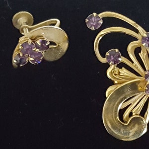 Puede incluir: Un conjunto de pendientes y un broche de tono dorado con diamantes de imitaci&oacute;n morados. Los pendientes son de estilo clip y el broche presenta un dise&ntilde;o floral.