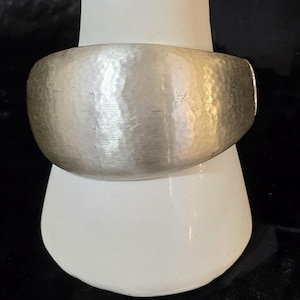 May include: A wide, silver-toned cuff bracelet with a hammered texture. The bracelet is displayed on a white, ceramic form against a dark background. The cuff is approximately 2 inches wide.
