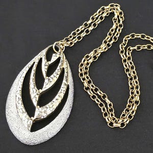 May include: A gold-tone necklace with a large, teardrop-shaped pendant. The pendant features a textured silver-tone outer layer and a gold-tone leaf design in the center. The chain is gold-tone with a lobster clasp.