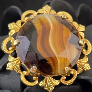 May include: A vintage brooch featuring a large, faceted, round gemstone with brown and amber tones. The gemstone is set in a gold-toned metal frame with ornate, leaf-like details and small decorative accents. The brooch has a classic, elegant design.