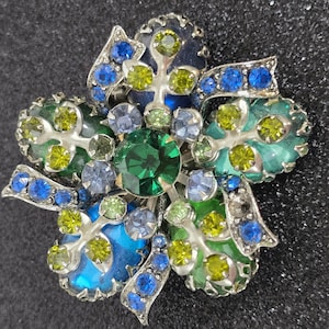May include: A floral brooch with a silver-tone metal base. The brooch features a central emerald-green stone surrounded by blue, green, and yellow gemstones. The petals are detailed with blue and green enamel.