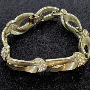 May include: A vintage gold-tone bracelet with a textured finish. The bracelet features a series of linked, stylized leaf-shaped elements, interspersed with sections adorned with small, clear rhinestones. The clasp is a fold-over style.