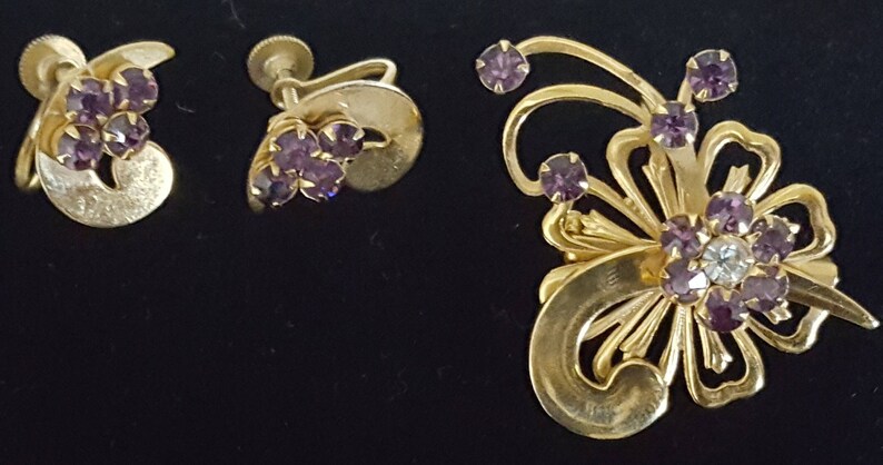 Puede incluir: Un conjunto de pendientes y un broche de tono dorado con diamantes de imitaci&oacute;n morados. Los pendientes son de estilo clip y el broche presenta un dise&ntilde;o floral.
