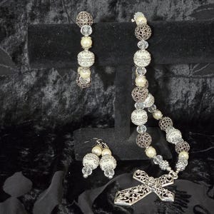 May include: A jewelry set featuring a bracelet, earrings, and a necklace with a cross pendant. The bracelet and earrings are displayed on a black velvet stand. The jewelry includes white, silver, and brown beads, and faux pearls.