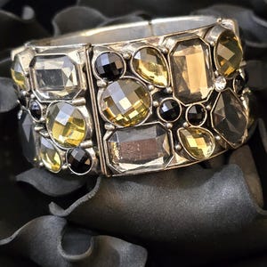 May include: A silver-toned bracelet with a mosaic of gemstones in various shapes and colors, including yellow, black, and smoky gray. The bracelet is set against a dark, textured background, highlighting the sparkle of the stones.