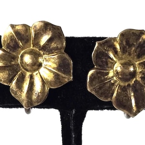May include: A pair of vintage clip-on earrings. Each earring features a gold-toned, flower-shaped design with detailed petals and a central, rounded element. The earrings are displayed against a black background.