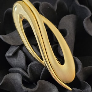 May include: A gold-toned metal brooch with an abstract, flowing design. The brooch has a smooth, polished surface and a curved, elongated shape. The jewelry piece is set against a dark, textured background.