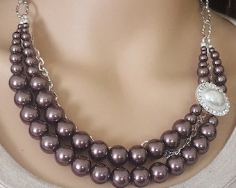 May include: A three-strand necklace featuring large, dark purple faux pearls. The necklace has a silver-tone chain and a decorative oval pendant with a large white pearl surrounded by clear crystals. The necklace is designed to be worn around the neck.