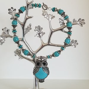 May include: A turquoise and silver owl necklace with turquoise beads. The necklace is displayed on a silver tree-shaped stand.